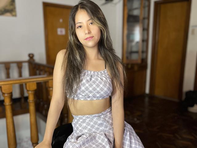 EvelinHotty, com cabelos castanhos longos, vestindo uma blusa e saia xadrez, sentada em uma cadeira de madeira, mostrando seus pequenos seios, em um quarto com iluminação quente..