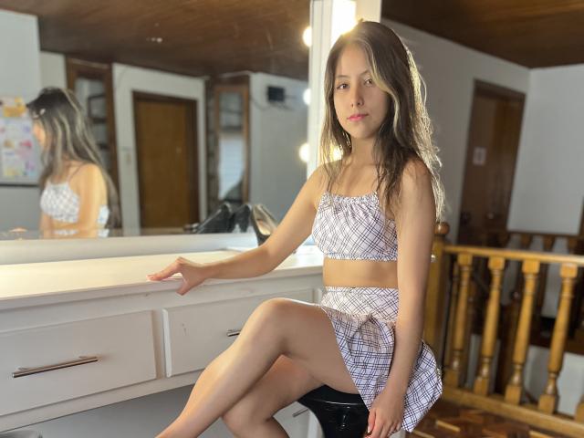 EvelinHotty, com cabelos castanhos ondulados, seios pequenos, e uma blusa cropped e saia xadrez, sentada em um banquinho preto em frente a um espelho em um quarto de madeira..
