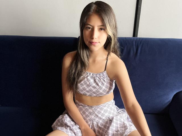 EvelinHotty mit langen, dunkelbraunen Haaren, die ein kariertes Crop-Top und einen Rock trägt, sitzt auf einem blauen Sofa und lächelt leicht in die Kamera. Ihre Brüste sind klein..