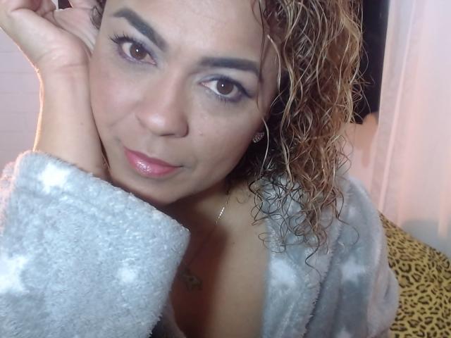 SolmaryHot with curly wet brown hair, wearing a fluffy gray robe, has brown eyes and glossy pink lips, touching her cheek with a hand, showing a cute smile.