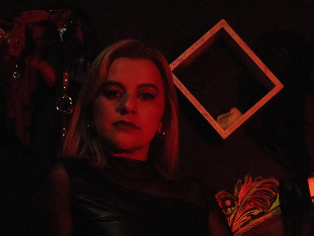 PerlaDivaio with red-lit face, blonde hair, black leather top, and colorful tattoo, looking slightly bored or annoyed. Empty frame in dark background.