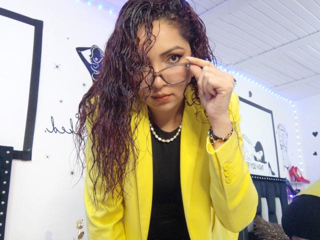Katrina Bello, com cabelo cacheado molhado, vestindo um blazer amarelo, uma blusa preta e um colar de pérolas, ajustando os óculos, olhando para a câmera, com um sorriso discreto, em um quarto bem iluminado..