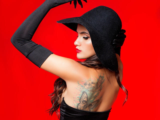 AmelieLautrec with black hat, long gloves, tattooed back, and red lipstick, showing off her ass and tits against a bright red background. She's looking sideways, raising one arm.