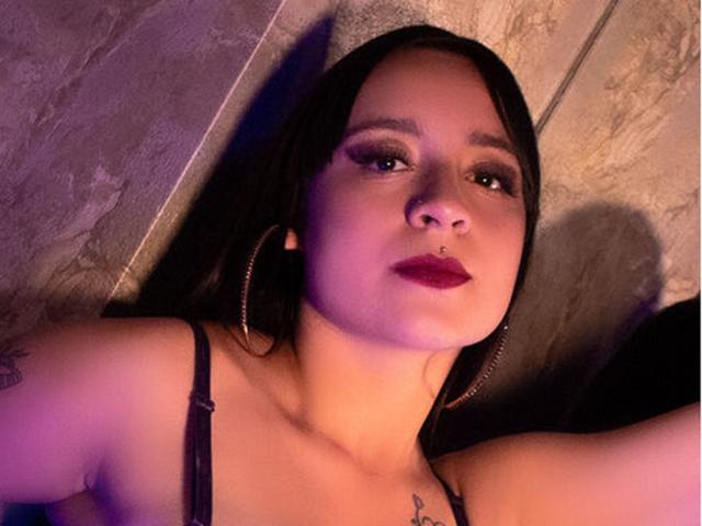 AnnPeeks tiene el pelo oscuro, aros grandes, un piercing en la nariz y lápiz labial rojo. Está mirando hacia arriba, con una iluminación morada y azul en su rostro, y tiene un tatuaje en su brazo izquierdo..