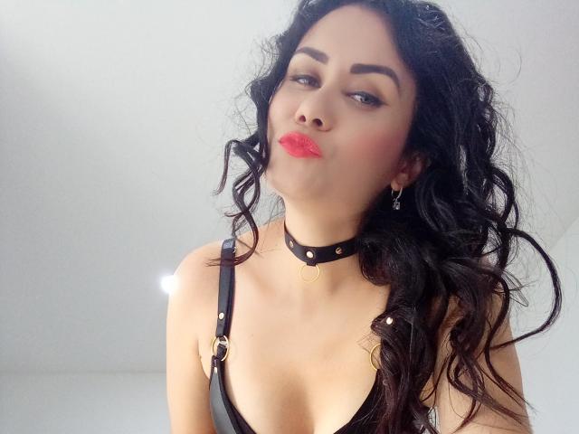 XBellaEvaX con cabello negro ondulado, vistiendo un collar negro y un vestido con tirantes, haciendo pucheros con lápiz labial rosa, mostrando sus pechos y mirando ligeramente hacia un lado en un selfie..