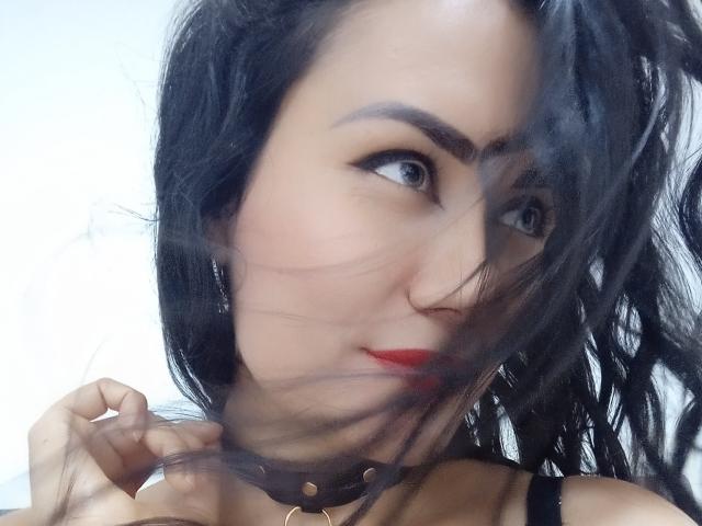 XBellaEvaX with black hair, red lipstick, and a black choker, her eyes looking sideways, with a wisp of hair across her face, in a slightly blurred selfie.
