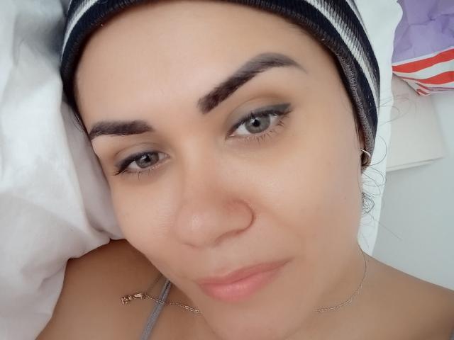 XBellaEvaX's face with light brown skin, green eyes, black eyebrows, subtle eyeliner, pink lips, small nose, wearing a striped headscarf, and a thin necklace, lying in bed.