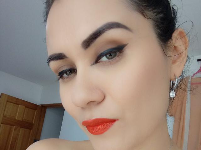 El rostro de XBellaEvaX, con lápiz labial rojo, delineador de ojos negro y cejas oscuras, lleva aretes de diamantes, mira ligeramente hacia abajo con ojos verdes y tiene una leve sonrisa, en una habitación luminosa..