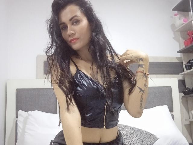 XBellaEvaX with long black hair, wearing a shiny black leather crop top, sitting on a bed, showing tattoos on her right arm, smiling with red lipstick.