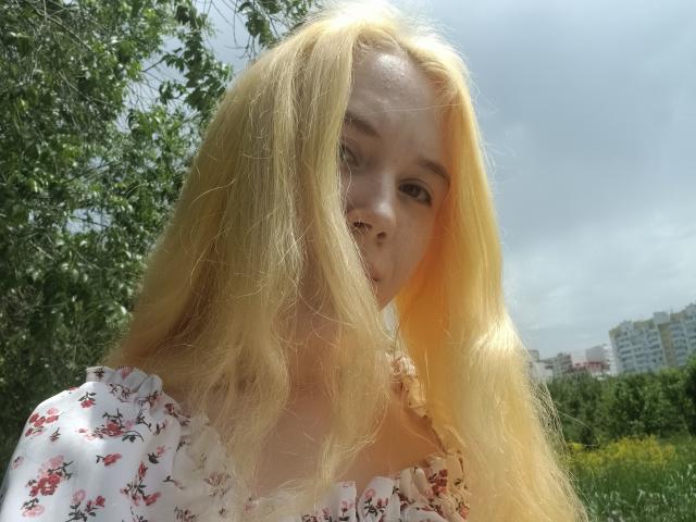 AnikaGlow with long, blonde hair, wearing a floral dress, has a nose ring and a slightly confused look, standing in a sunny, green, outdoor setting.
