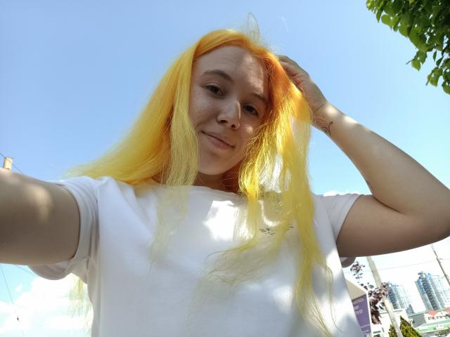 Anika, con cabello largo y rubio, una camiseta blanca y piel clara, sonríe mirando hacia la cámara, con una mano en el cabello. Está afuera en un día soleado..