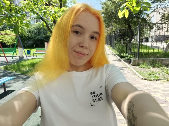 AnikaGlow with bright yellow hair, white t-shirt, small tits, fair skin, freckles, smiling, outdoor selfie, holding phone, tattoo on left forearm, sunny park, green trees.