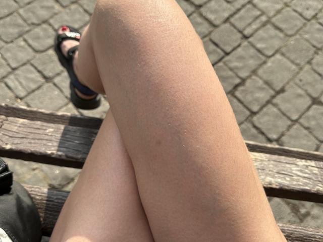 Las piernas de BellaJulls estaban cruzadas sobre un banco de madera, mostrando una piel lisa y bronceada, con una ligera línea de bronceado, y llevaba chanclas negras con tiras rojas..