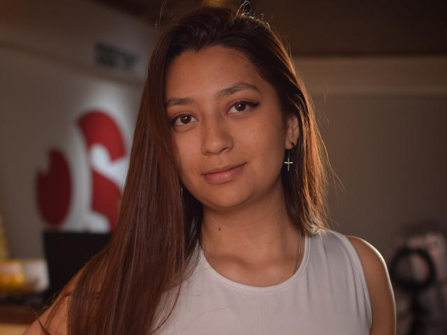 VioletaBlair, avec de longs cheveux bruns, de petites boucles d'oreilles en forme de croix, et un haut blanc sans manches, sourit doucement à la caméra, montrant un visage mignon et séduisant..
