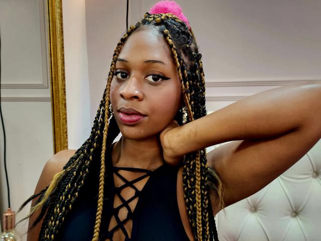 KarmenCastillo with braided hair, pink pom-pom, black lace-up top, arm raised, looking at camera, medium brown skin, pink lips, gold earrings, sexy, confident, cute.