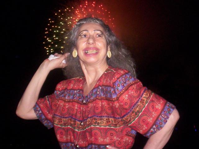 ElenahHottie met grijs haar, die een rood, geruit shirt draagt, gele oorbellen en een witte ring, lachend met haar hand op haar heup, vuurwerk op de donkere achtergrond..