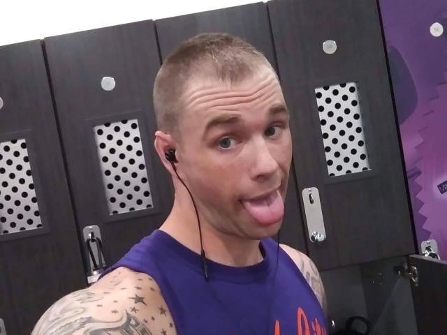 JetJhonson, con la cabeza afeitada, tatuajes en los brazos, una camiseta morada sin mangas, auriculares negros, la lengua afuera, mirando a la cámara, en un vestuario con armarios de madera oscura..