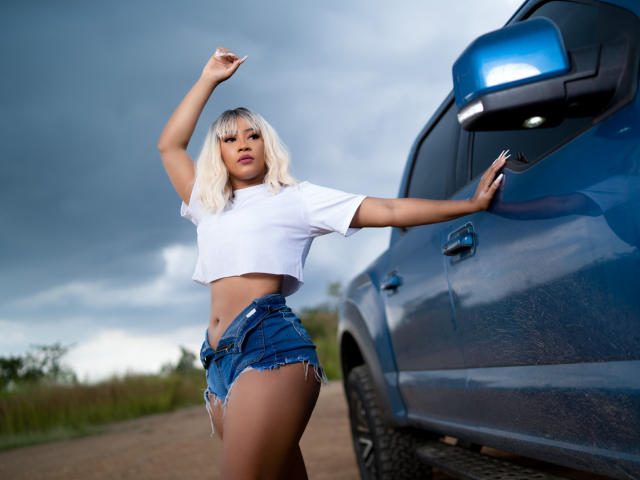 Mariam Lowel, con cabello rubio, un top blanco corto y pantalones vaqueros cortos deshilachados, apoyada en un coche azul, con el brazo levantado, bajo un cielo nublado, mostrando su trasero y una expresión de confianza..