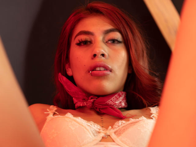 DalylaLove a les cheveux roux, un bandana rose et un soutien-gorge en dentelle blanche, la langue tirée, les yeux levés avec du crayon noir, ses seins sont visibles, lumière douce et la bouche légèrement ouverte..