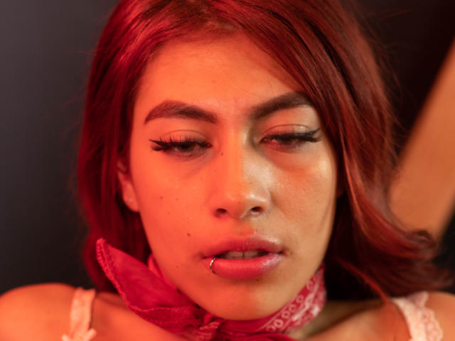 DalylaLove met rood haar, haar tong uit, draagt een roze bandana, heeft dikke wimpers, een neusring en kijkt naar beneden met licht geopende lippen. Rode belichting..