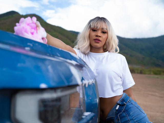 Mariam Lowel, con i capelli biondi, una canotta bianca e jeans blu, sta lavando un'auto blu con sapone rosa, con la mano sul cofano dell'auto, e montagne sullo sfondo..