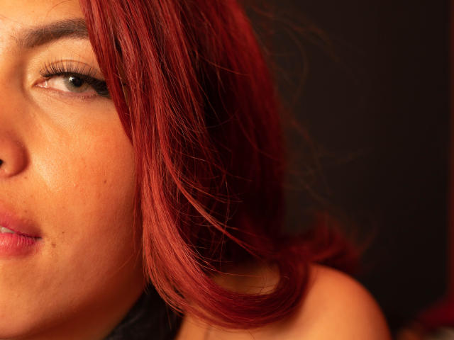 Le visage de DalylaLove, avec ses cheveux roux, ses yeux verts et ses lèvres légèrement entrouvertes de couleur rose, révèle une petite tache de rousseur sur sa joue. Sa peau est de couleur chaude et présente des taches de rousseur visibles..