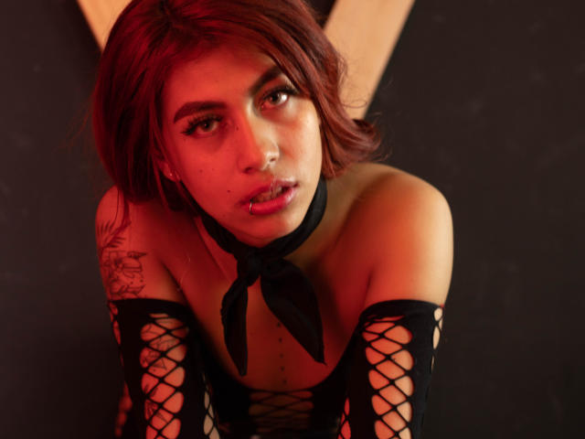 DalylaLove, avec des cheveux roux, des manches en résille noire, un foulard noir et un tatouage sur le bras gauche, regardant vers le haut avec les lèvres entrouvertes. Éclairage rouge et orange chaud. Expression douce et séduisante..