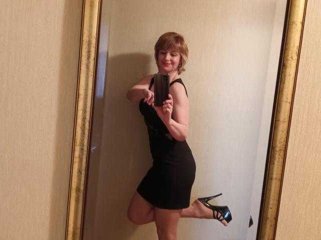 NatashaNannette in a black dress, holding a phone, smiling, with short red hair, black high heels, and her ass slightly raised in a mirror selfie.