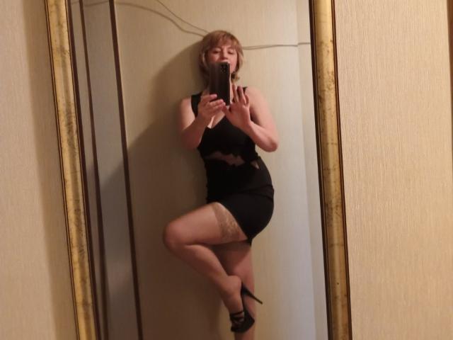 NatashaNannette with blonde hair, black dress, and thigh-high stockings, standing on one leg, taking a mirror selfie with her phone, showing her ass and tits.