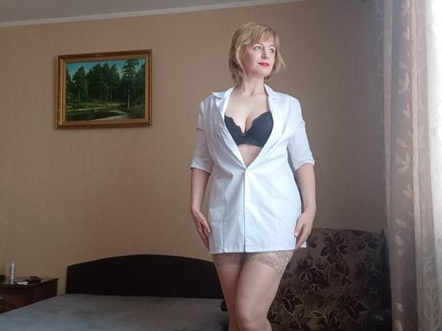 NatashaNannette with short blonde hair, wearing a white button-up shirt open to reveal black bra and lace garters, standing in a bedroom, showing her tits and ass.