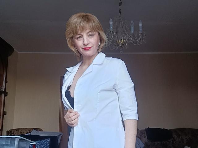 NatashaNannette, blonde avec des cheveux courts, portant une chemise blanche, laissant apparaître un soutien-gorge noir d'une main, avec du rouge à lèvres, se tient dans une pièce où se trouve un lustre en cristal au-dessus..