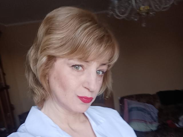 NatashaNannette has short blonde hair, green eyes, and red lipstick. She's wearing a white shirt, looking slightly to the side with a subtle smile. Chandelier in the background.