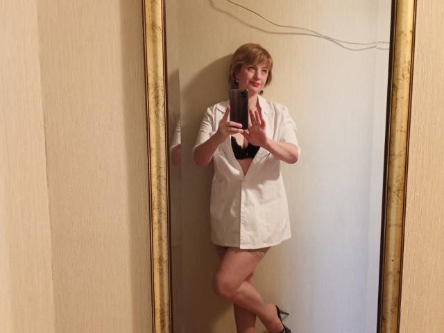 NatashaNannette, avec ses cheveux blonds courts, porte une chemise blanche déboutonnée, un soutien-gorge noir et des talons noirs, et prend un selfie devant un miroir, montrant ses seins et ses fesses, debout avec une jambe croisée..