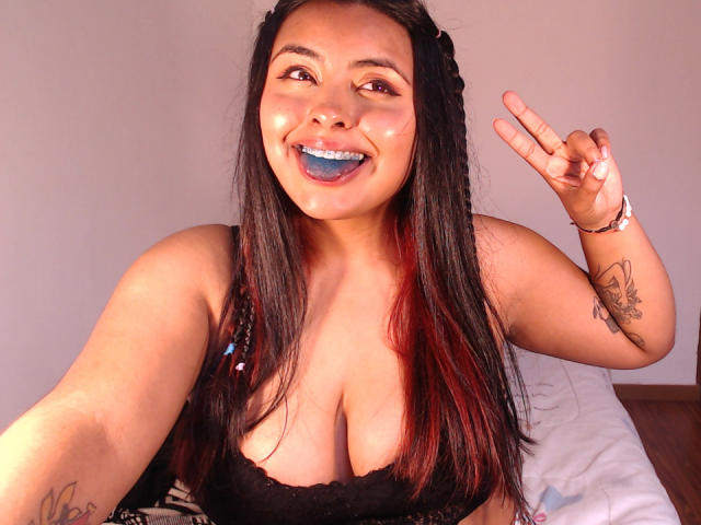 AngelaRoggers with blue lipstick, black lace bra, showing big tits, smiling, long braided hair, tattoo on right arm, flashing peace sign, tan skin, brown eyes, wood floor.