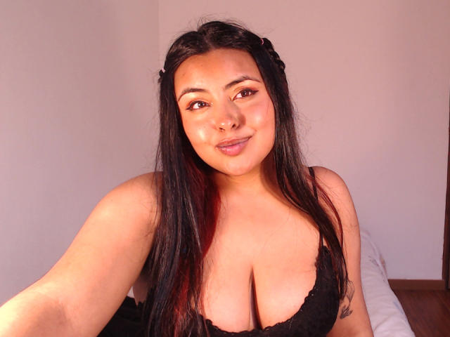 Angela Roggers, con cabello negro largo, vistiendo un sujetador de encaje negro, mostrando sus grandes senos, sonriendo a la cámara, con luz solar sobre su piel bronceada, en un dormitorio..