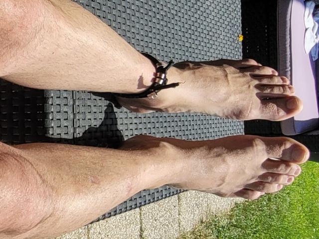 Le gambe pelose di Oli69, con una cavigliera in pelle marrone, piedi nudi e suole leggermente sporche, sono distese su un tappetino nero con una texture, con dell'erba nell'angolo in basso a destra..