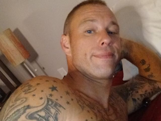 JetJhonson sin camisa, con tatuajes, ojos azules, cabello rubio corto y una sonrisa, apoyado en su brazo. Se ven tatuajes en los hombros y el brazo, acostado en una cama..