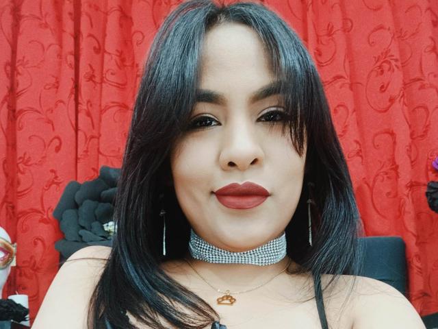 SharomCastle with long black hair, wearing red lipstick, a sparkly choker, and earrings, smiling at the camera, showing her face and upper body. Boobs not visible.