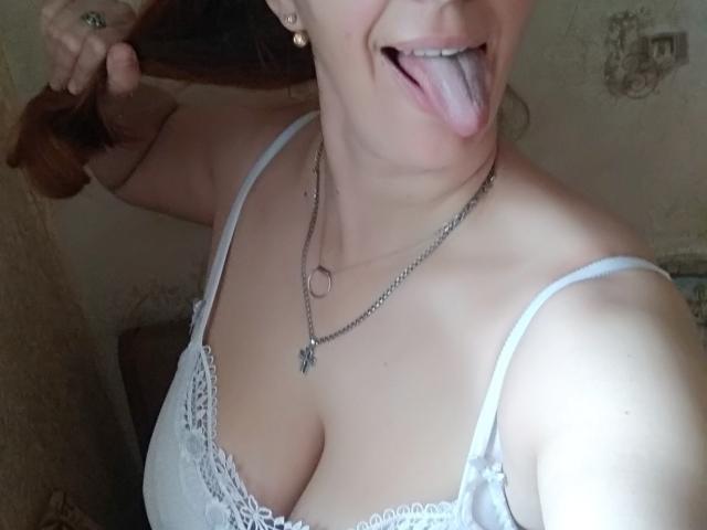 MissieLorrie with her tongue out, holding her brown hair, wearing a white lace bra, showing her tits, and a necklace with a ring and chain.