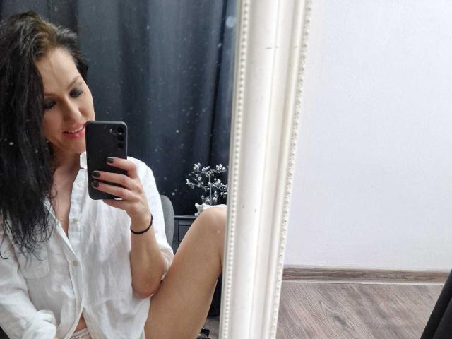 JoliDanielle with black hair, wearing a white button-up shirt, sitting in front of a mirror, taking a selfie with her phone, showing her thigh and smiling.