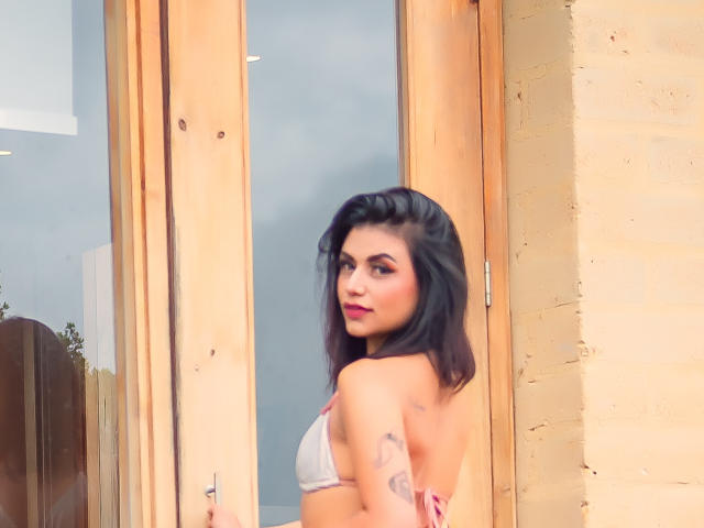 Kristal Jacobs, con i capelli scuri, guarda indietro, indossa un reggiseno rosa e bianco, mostrando il seno e i tatuaggi sul braccio destro, ed è in piedi vicino a una porta di legno..
