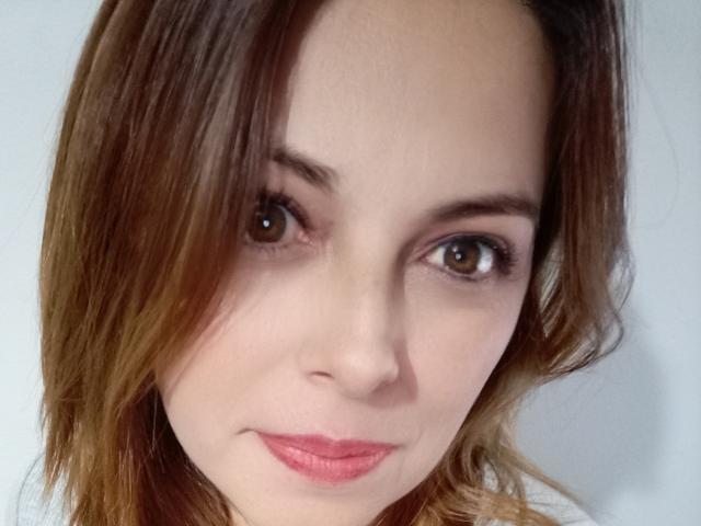 Seher's face, with brown eyes, light skin, and pink lipstick, has brown, wavy hair covering one eye, smiling slightly. Her tits are not visible.