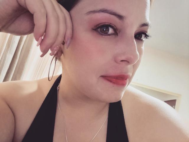 O rosto de Seher, com sombra rosa, batom vermelho, brincos de argola e um top preto de alças finas. Ela está apoiando a cabeça na mão, exibindo suas unhas bem cuidadas. Iluminação suave..
