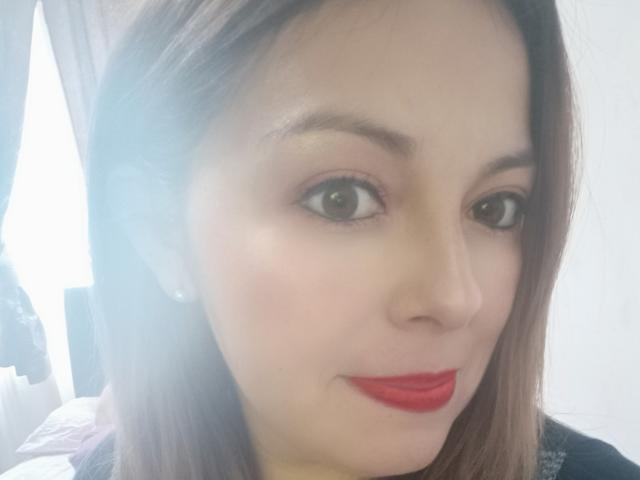 Seher's face with light skin, brown eyes, red lipstick, and straight brown hair, wearing small earrings, in a soft light. She's smiling subtly.
