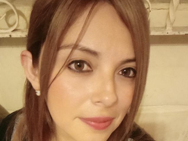 Seher's face with brown eyes, light skin, and pink lipstick, wearing small diamond earrings, has straight brown hair, and a slight smile, looking at the camera.