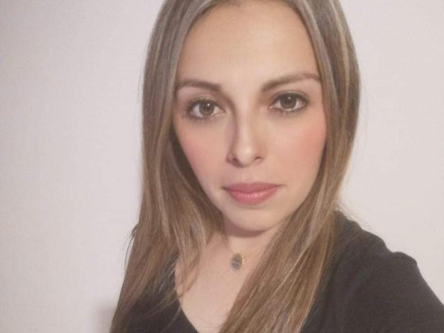 Seher has long brown hair, brown eyes, pink lips, and a slight blush. She's wearing a black top and a small necklace with a tiny pendant.