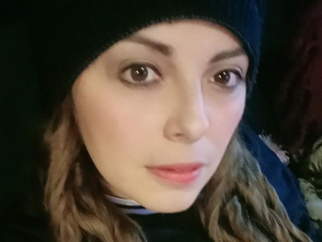 Seher's face, wearing a dark knit hat, with wavy brown hair, light skin, and brown eyes, has pink lips and a slightly smiling expression. Boobs partially visible under hat.