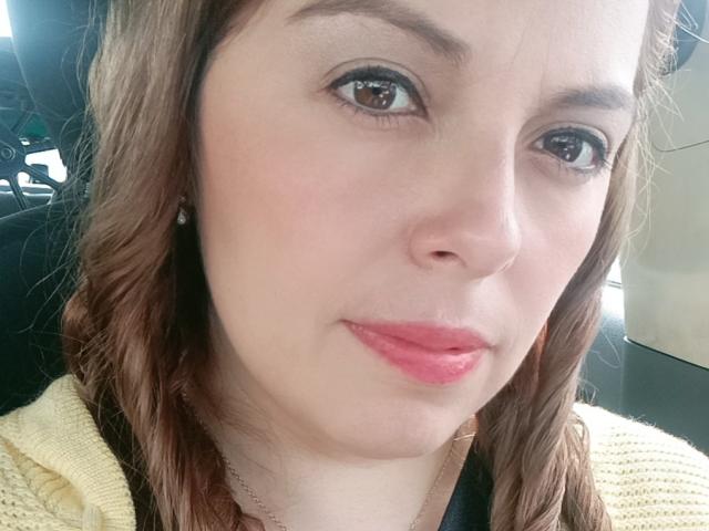 El rostro de Seher, piel clara, ojos marrones, lápiz labial rosa, labios ligeramente fruncidos, cabello ondulado castaño, suéter amarillo, aretes pequeños, mirando a la cámara, expresión suave, ligera sonrisa, maquillaje sutil..