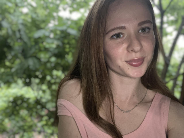 HereIsSoul with long brown hair, pink top, small nose, subtle smile, light skin, and a thin necklace, looking at the camera with green eyes in a natural outdoor setting.