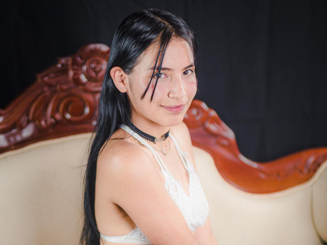 TiffanyEve mit schwarzen Haaren, weißem Spitzen-BH, schwarzem Choker, lächelnd, sitzend auf einem verzierten Holzstuhl, die Brüste zeigen, süß, helle Haut, asiatisch, weiches Licht, entspannte Pose..